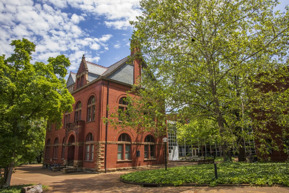 A red brick building with arched windows and a steep, gabled roof, partially obscured by large, leafy green trees. The building sits on a paved path surrounded by lush greenery, with a bright blue sky and scattered clouds overhead.